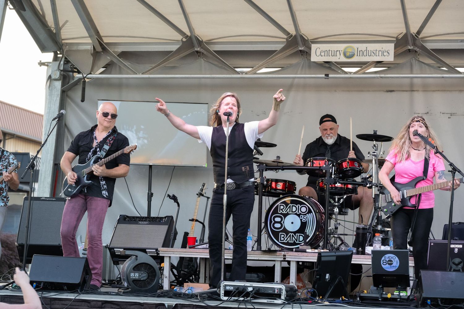 Band members performing on an outdoor stage with instruments and Radio 801 drum kit.