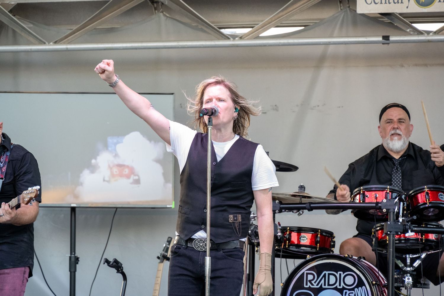 A rock band performs energetically on stage with a lead vocalist and drummer visible under a canopy.