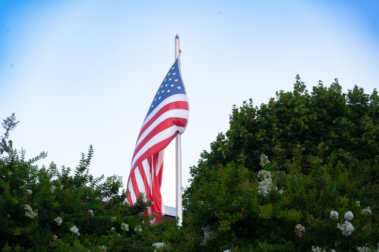 American flag waving against blue sky with green trees in background.