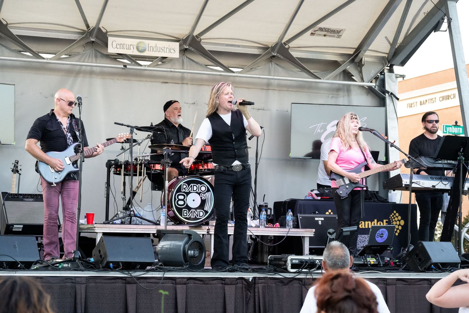 A live band performs on an outdoor stage with instruments including guitar, drums, and keyboards.