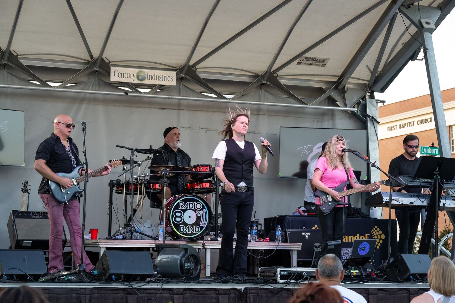 Band playing energetically on a covered outdoor festival stage during a daytime performance.