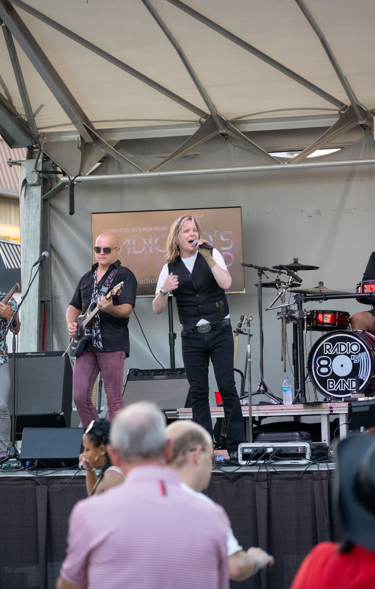 Musicians performing under a white tent canopy at an outdoor event with crowd watching below.