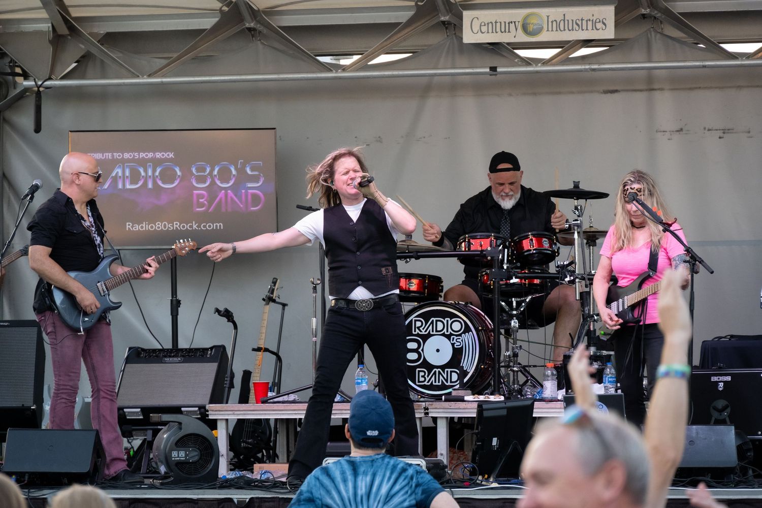 Band members playing instruments and singing on stage during an outdoor concert performance.