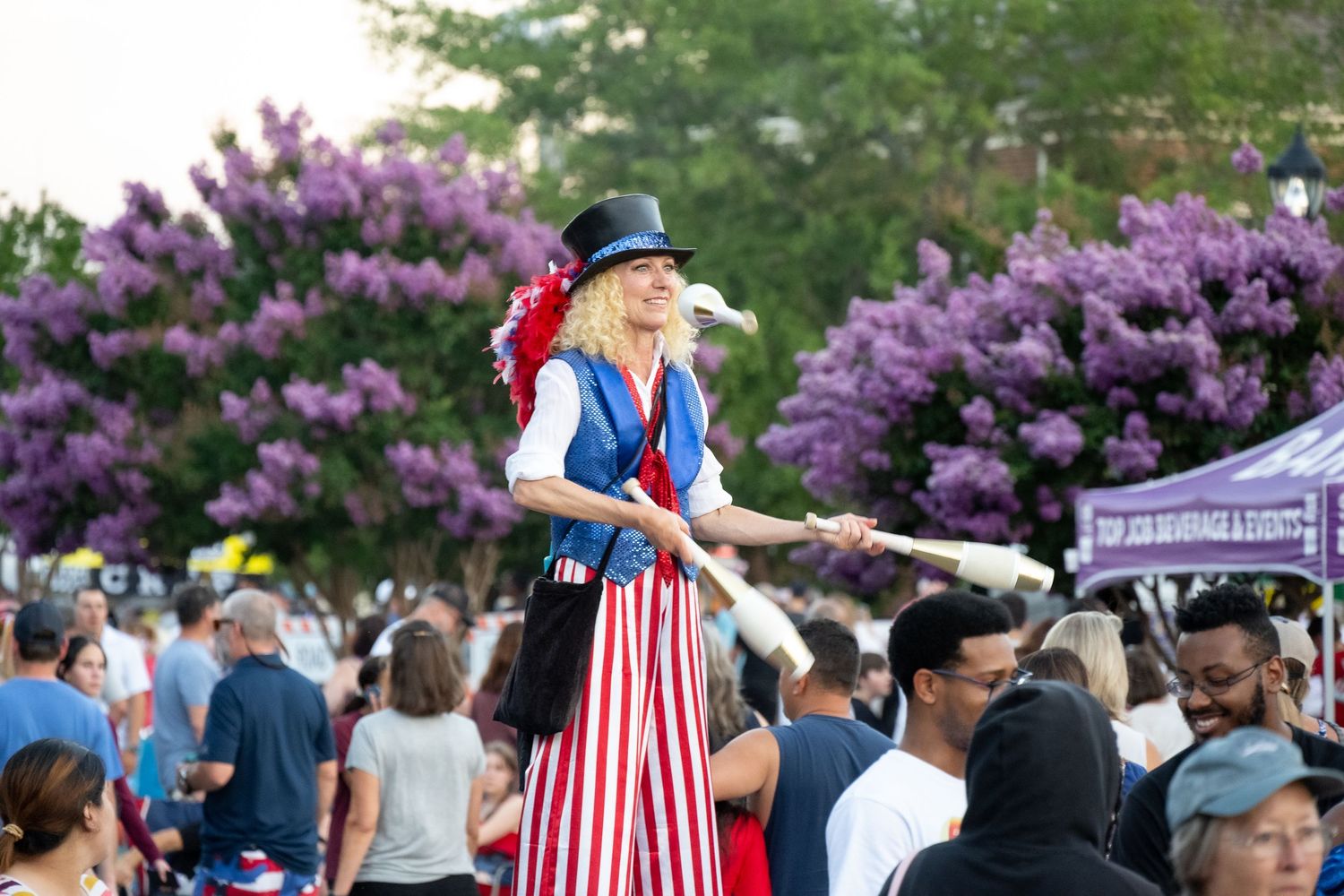 Performer in patriotic attire entertaining crowds at an outdoor summer festival with purple flowers in background.