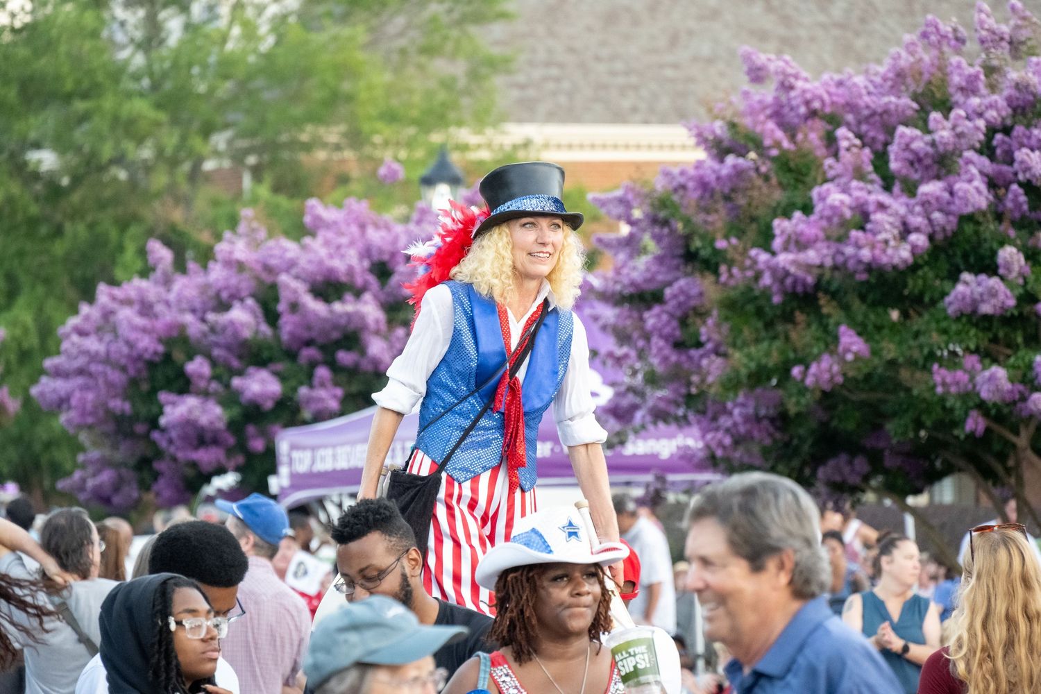 A person in patriotic Uncle Sam style costume entertains a crowd near blooming purple lilac bushes.
