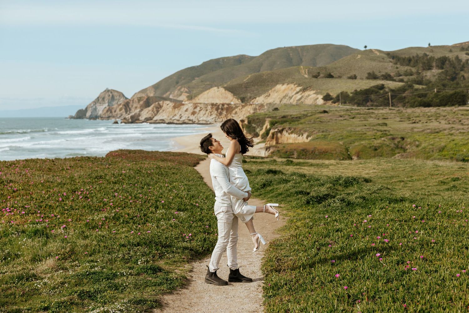 a man holding his fiance up on a cliffside in Pacifica, California during their proposal and engagement photoshoot in the Bay Area