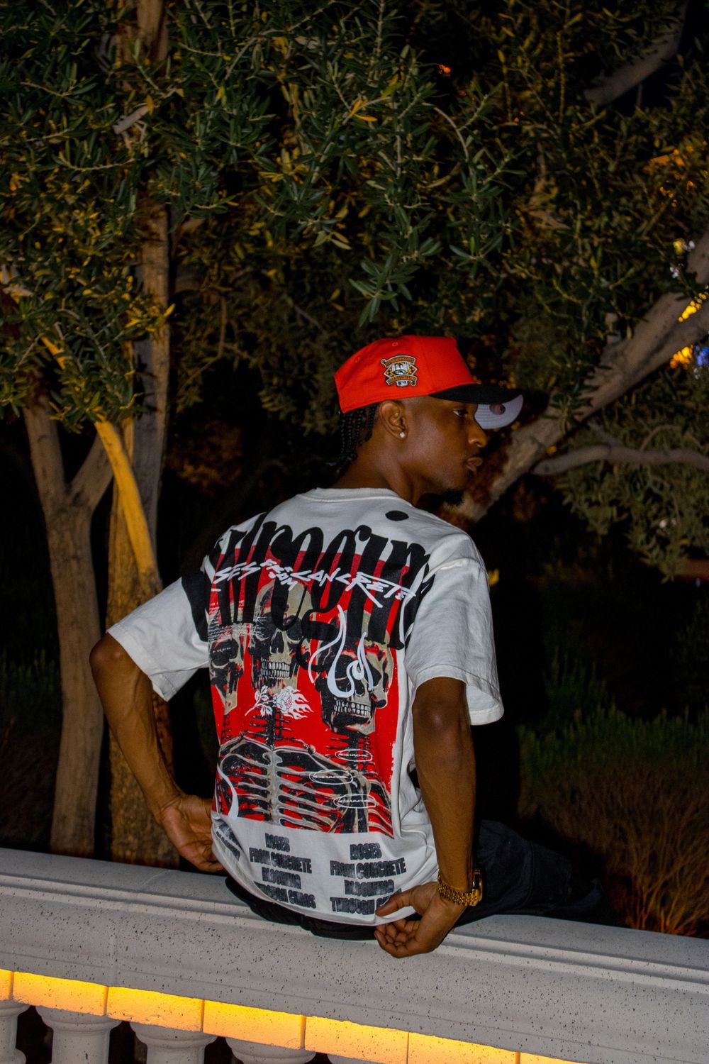 Young man in red cap and graphic tee poses by balcony railing at night.