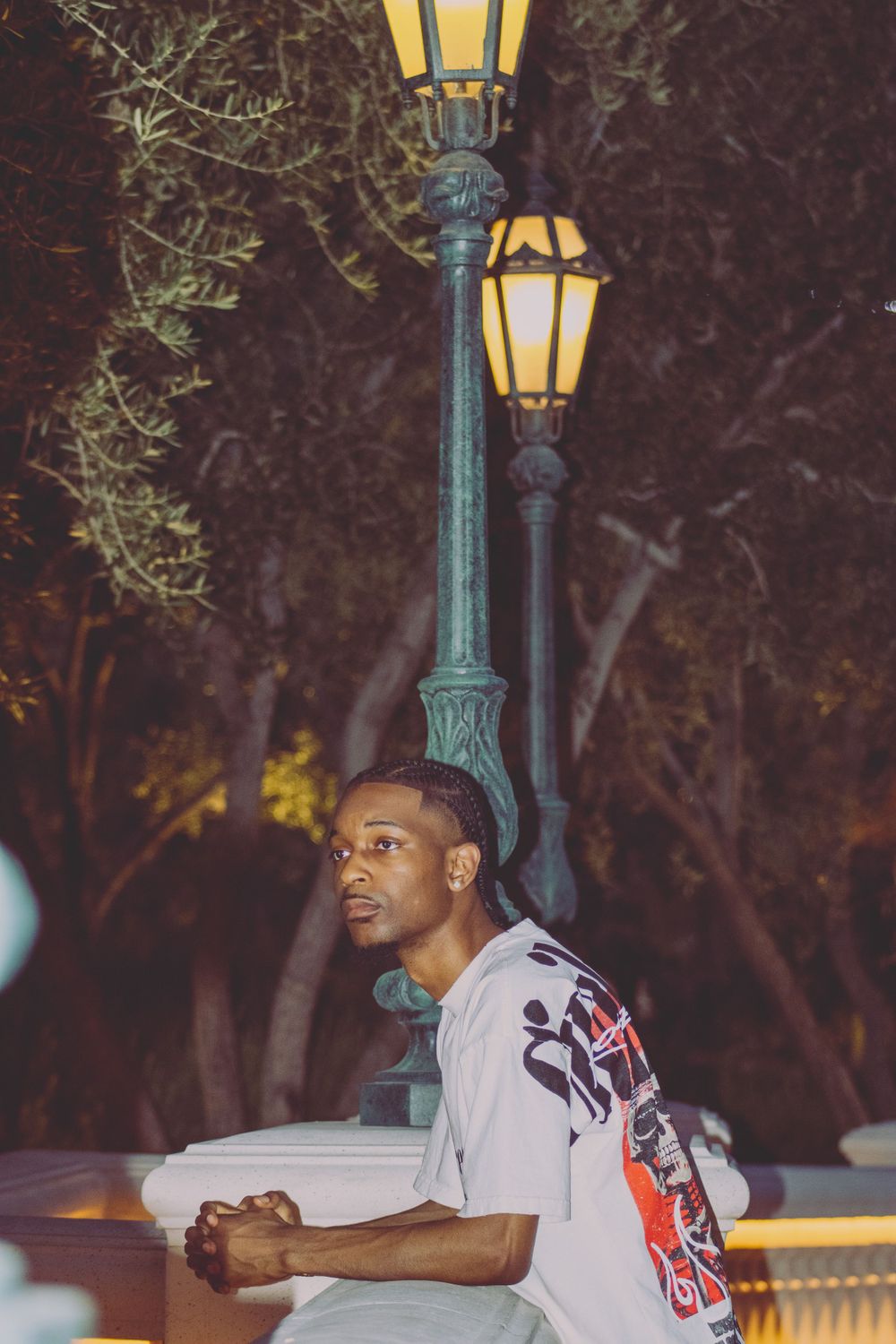 Young Black man in graphic tee leaning on wall near vintage street lamp at night.