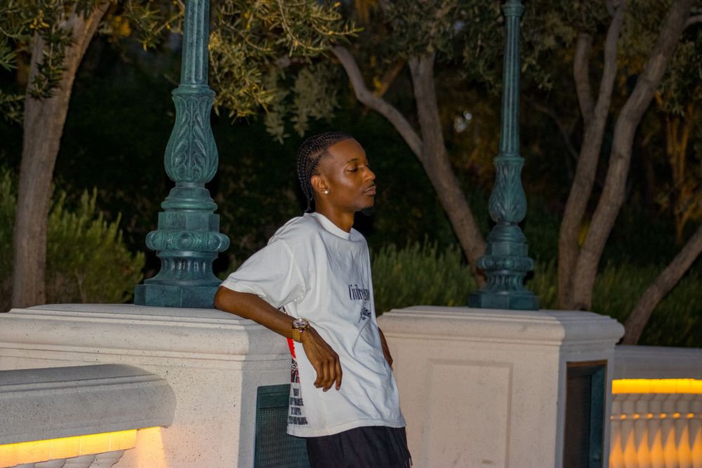 Young man in white oversized tee leans against stone wall outdoors at night.