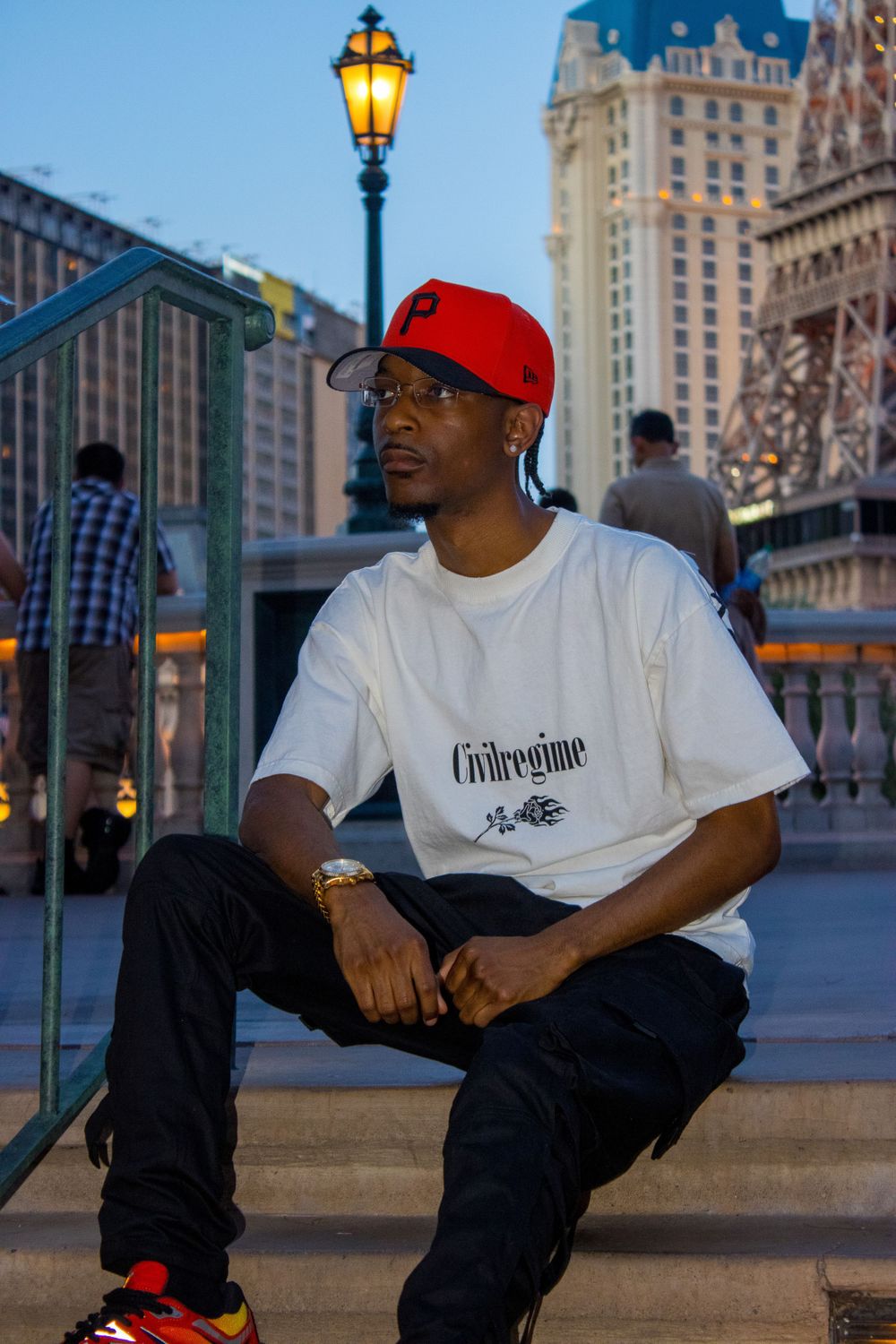 Man in red cap and Civilregime tee sits on steps near Paris Las Vegas at dusk.