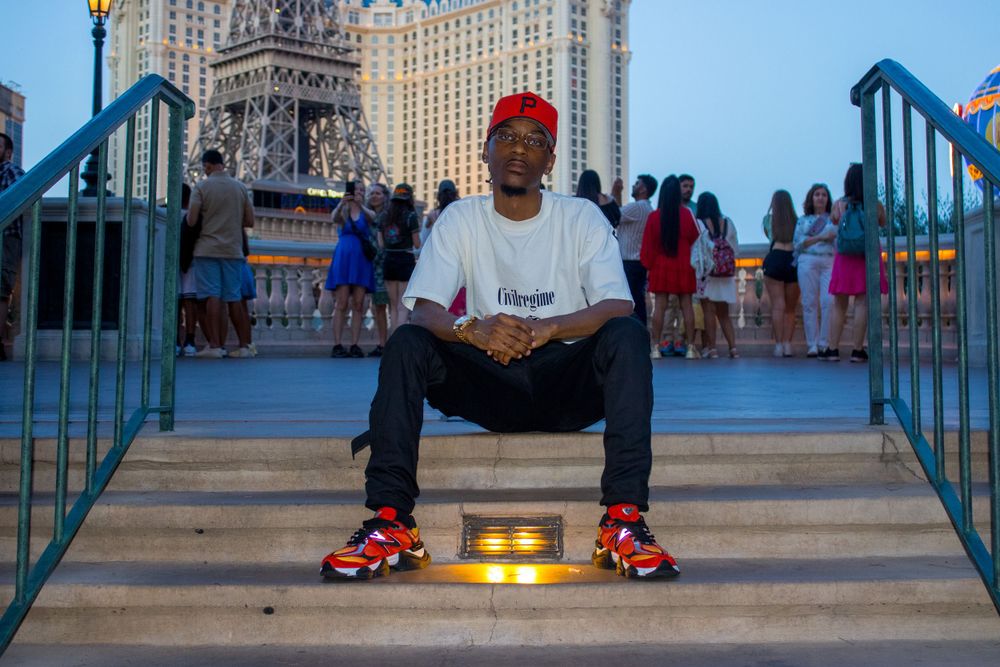 Young man in white tee, red cap, and colorful sneakers sitting on steps near Las Vegas Eiffel Tower replica.