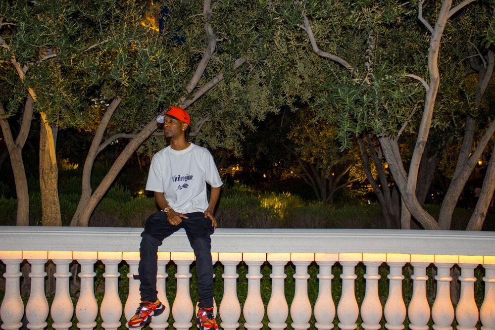 Young man in orange cap and white tee sits on white balustrade surrounded by trees at night.