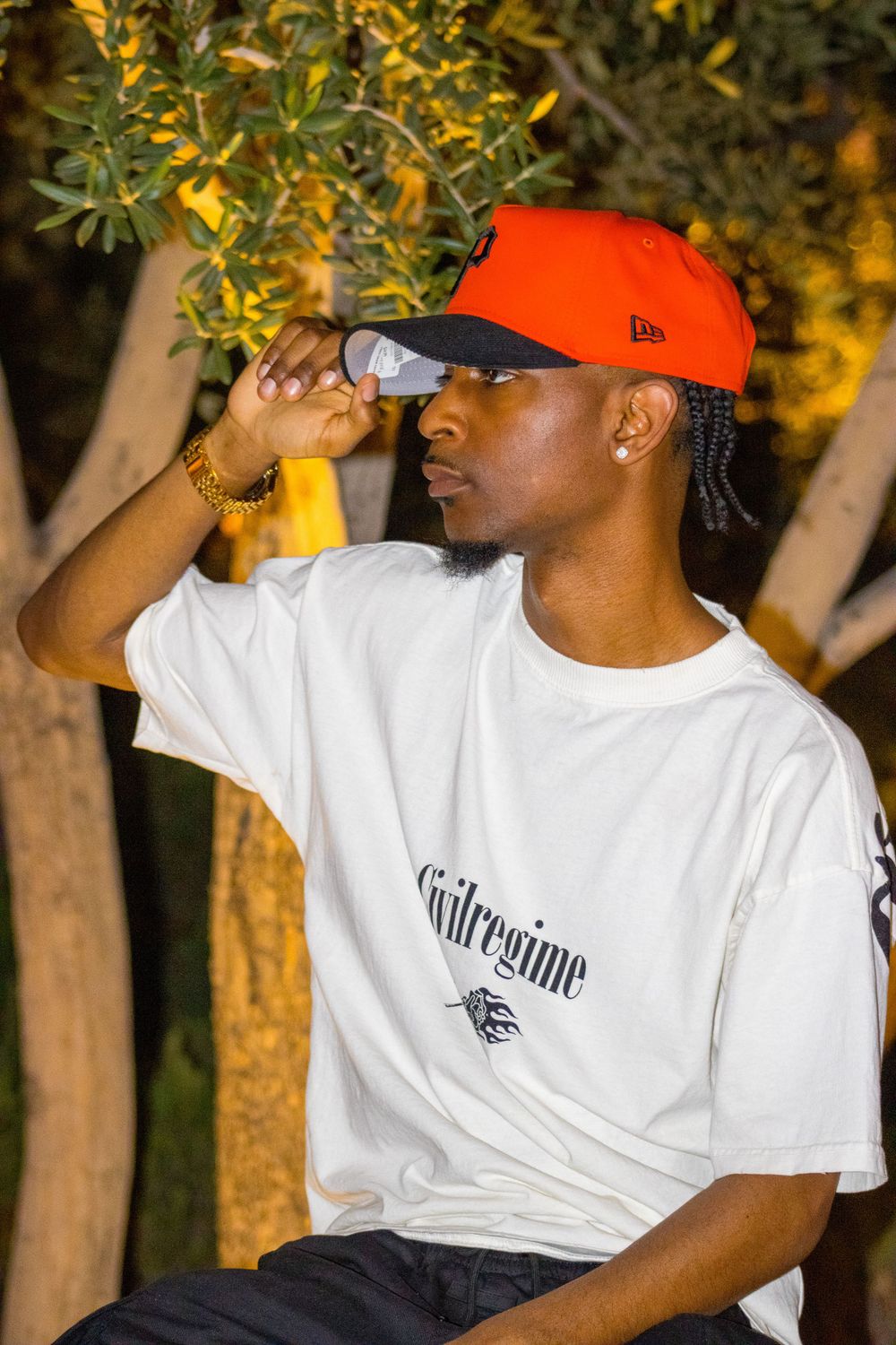 Young man in red New Era cap adjusts brim while wearing white Civilregime tee outdoors.