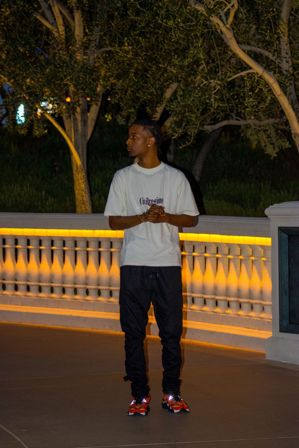 Young man in white tee and black joggers standing near illuminated balustrade surrounded by trees at night.