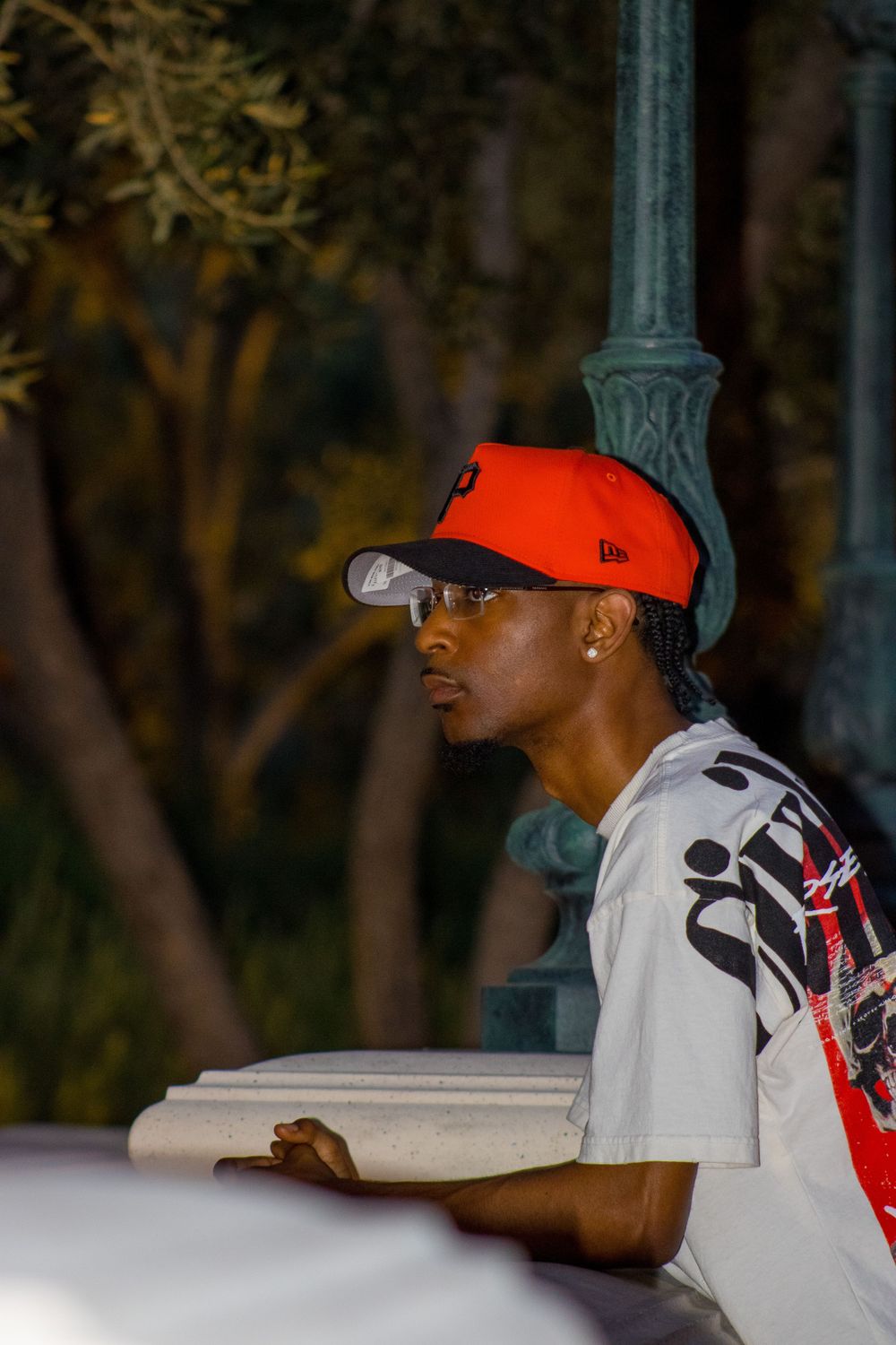 Young Black man in orange New Era cap and white graphic tee leaning on white railing near green lamp post.