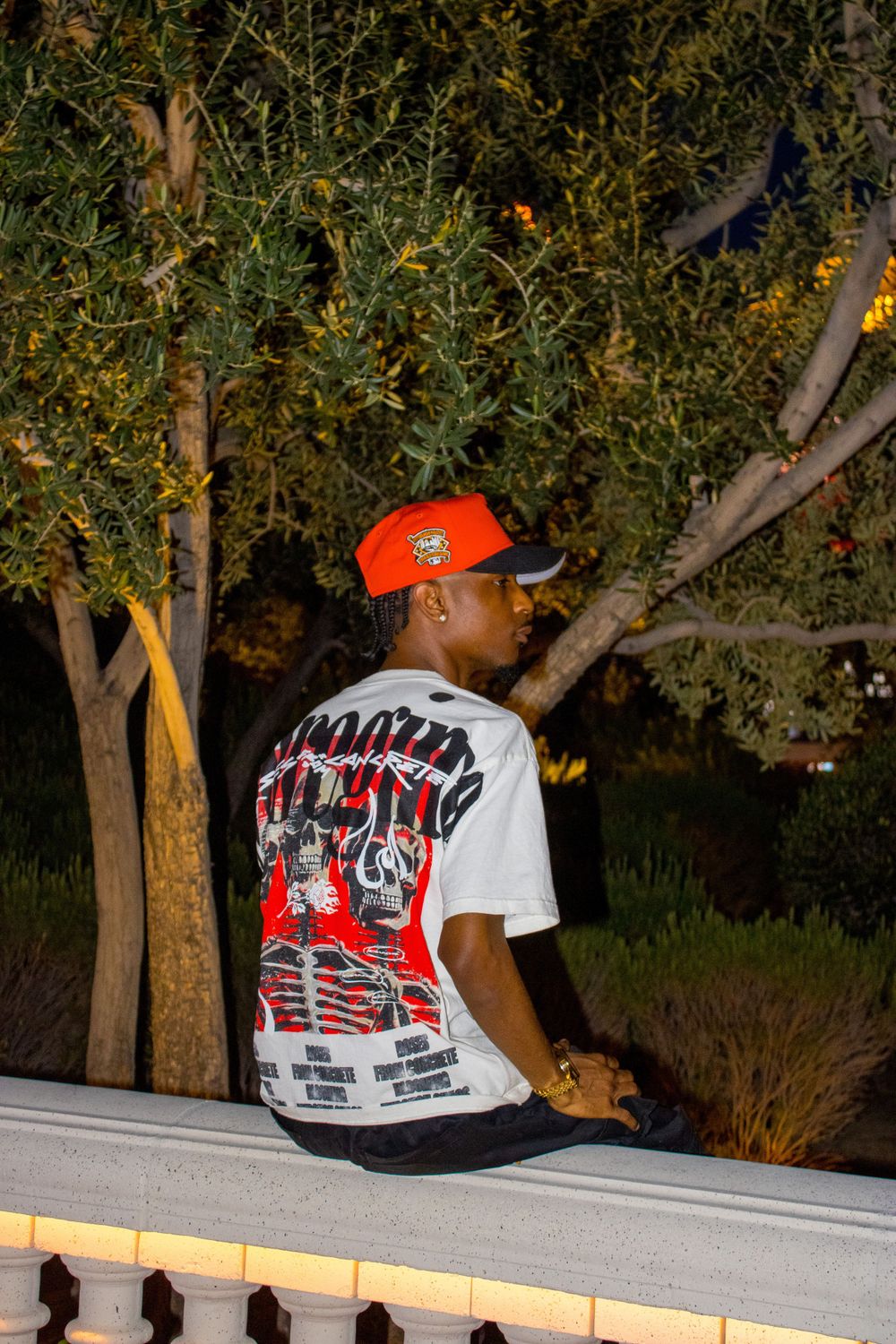 Young Black man in orange cap and white graphic tee sitting on balustrade with trees at dusk.