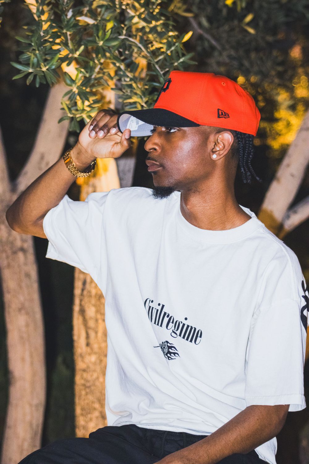 Young Black man in orange New Era cap and white Civil Regime tee adjusting hat brim outdoors.