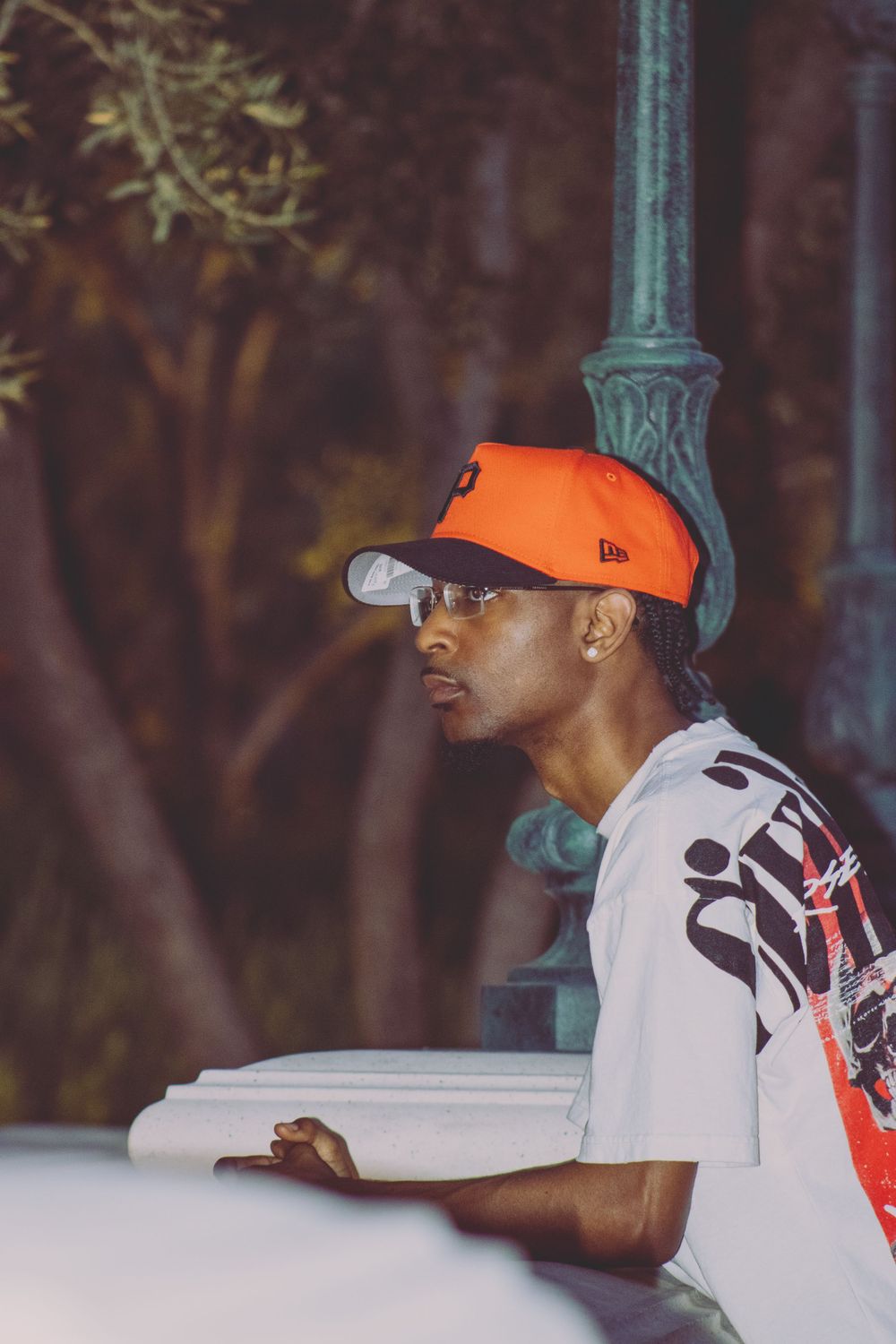 Young Black man in orange snapback and white graphic tee leaning on white railing near lamp post.