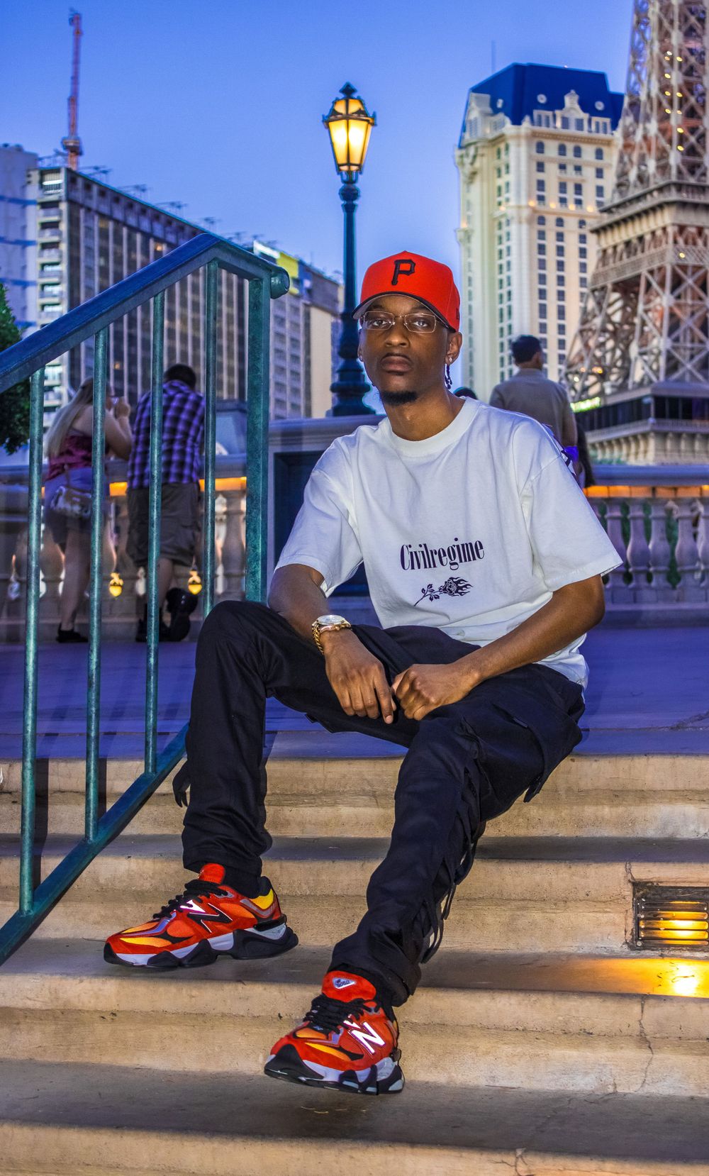 Young Black man in red P cap, white Civil Regime tee, and New Balance sneakers posing on steps.