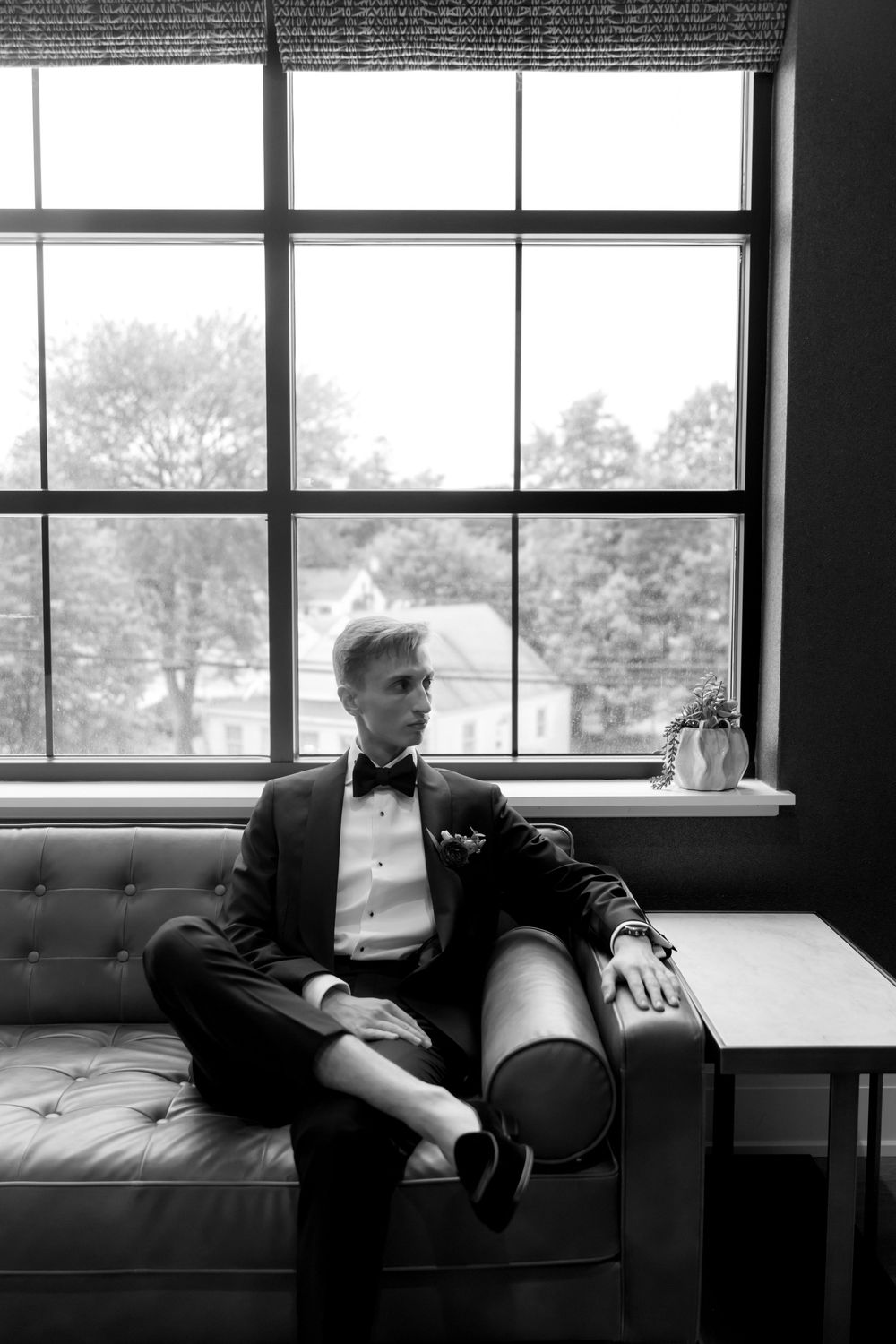 Moody black and white portrait on a leather couch by large windows.
