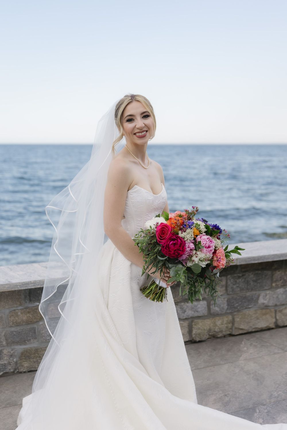 A bride in a white dress holds a vibrant bouquet of red and pink flowers against an oceanfront backdrop.