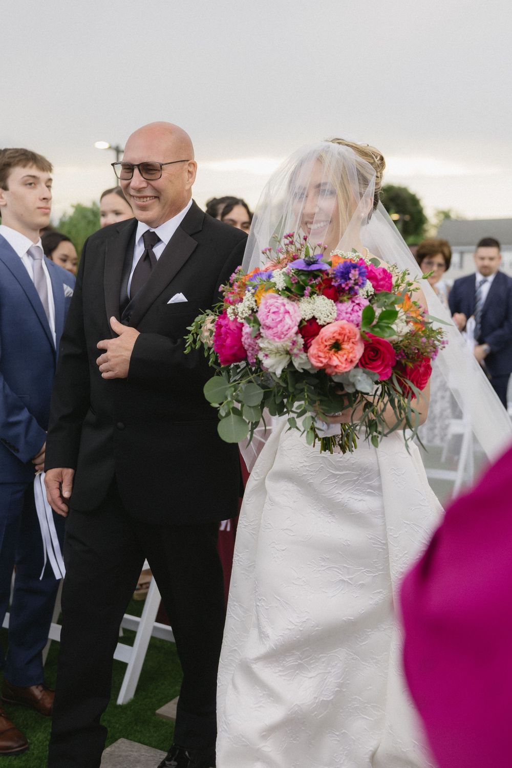 Wedding party gathers during ceremony with bright floral arrangements and white decor.