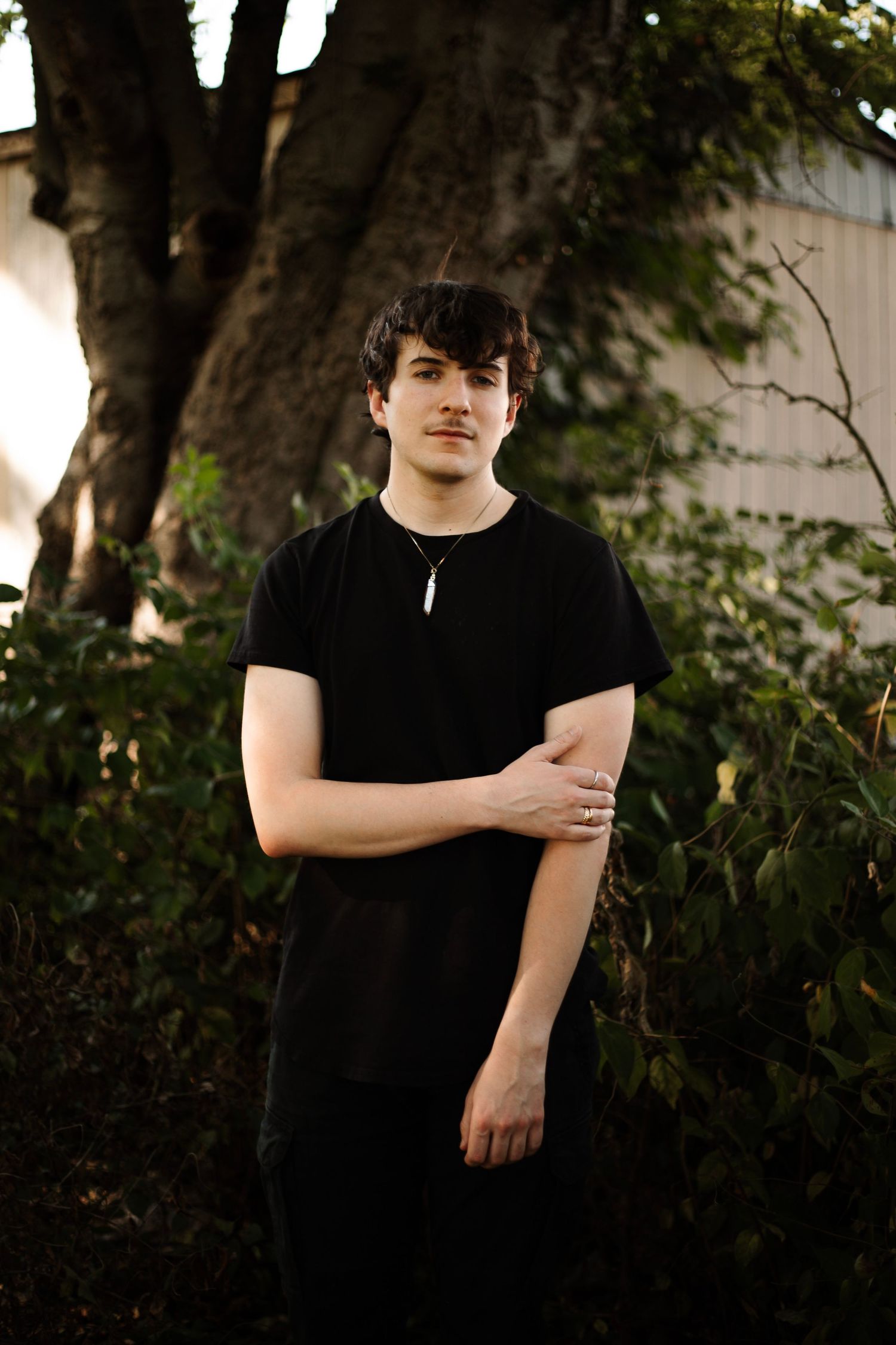 Young person in a black t-shirt stands outdoors against foliage in warm evening light.