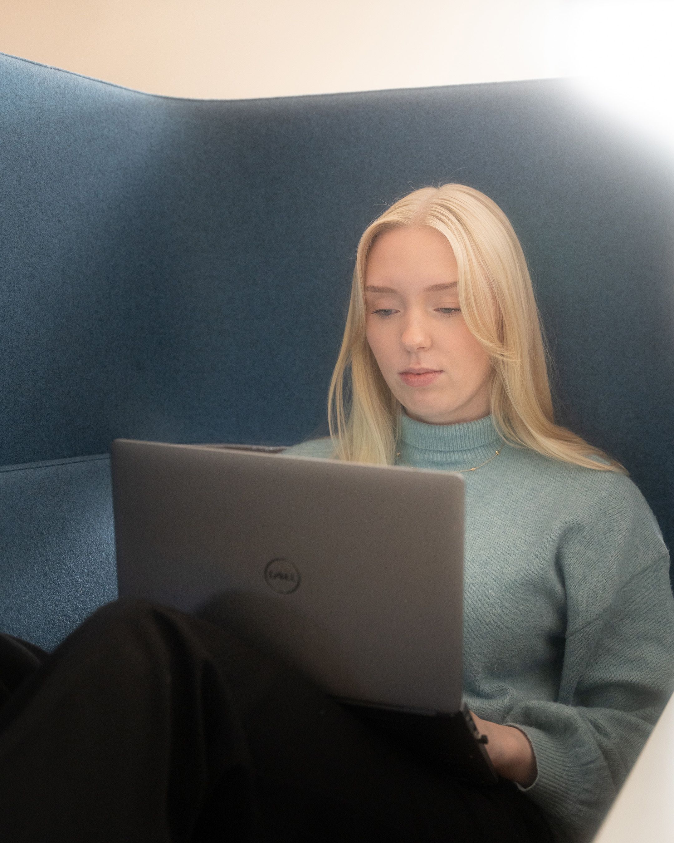 Person in a grey sweater working on a Dell laptop while sitting on a blue couch.