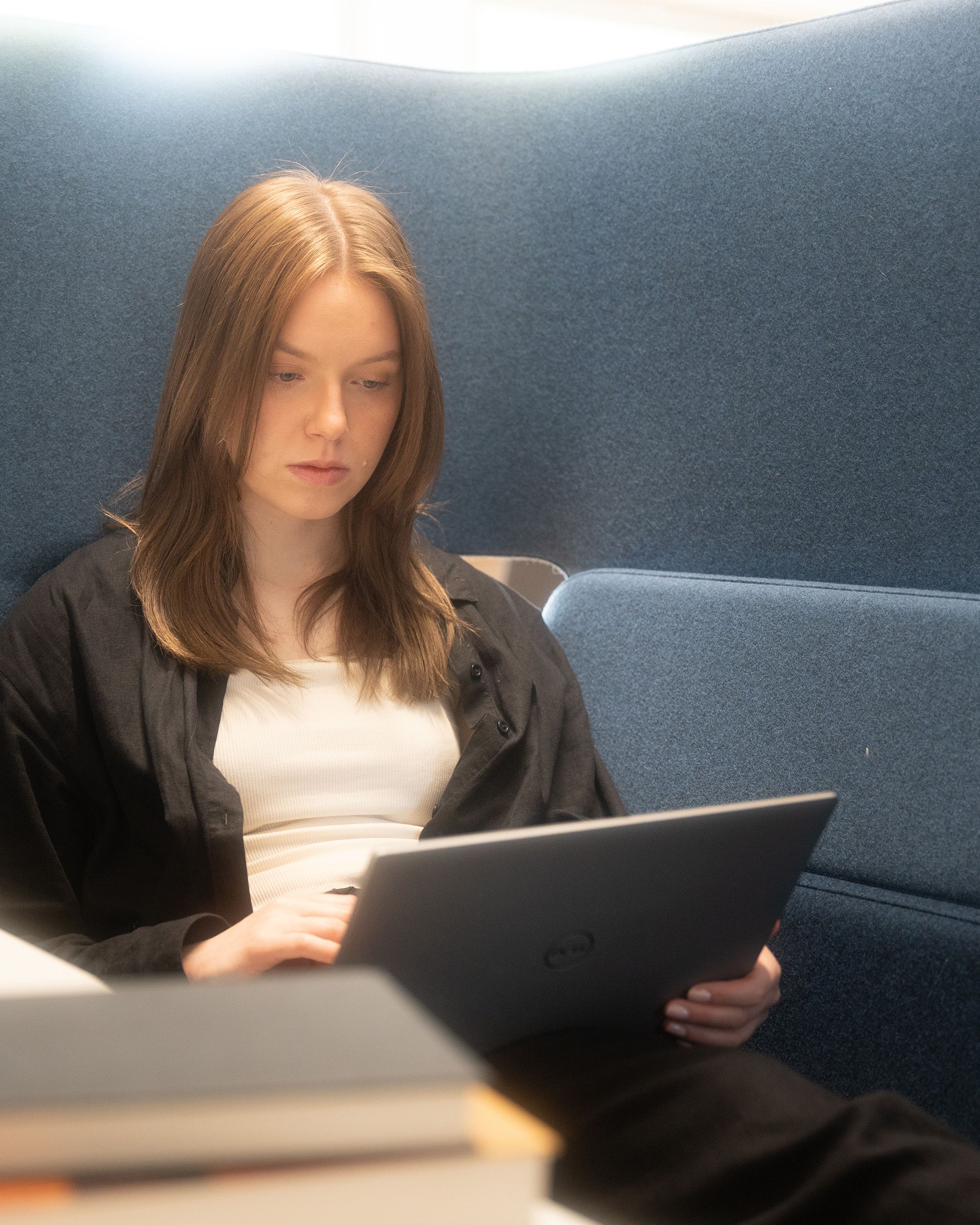 Person working on a laptop while sitting on a blue sofa in dim lighting.
