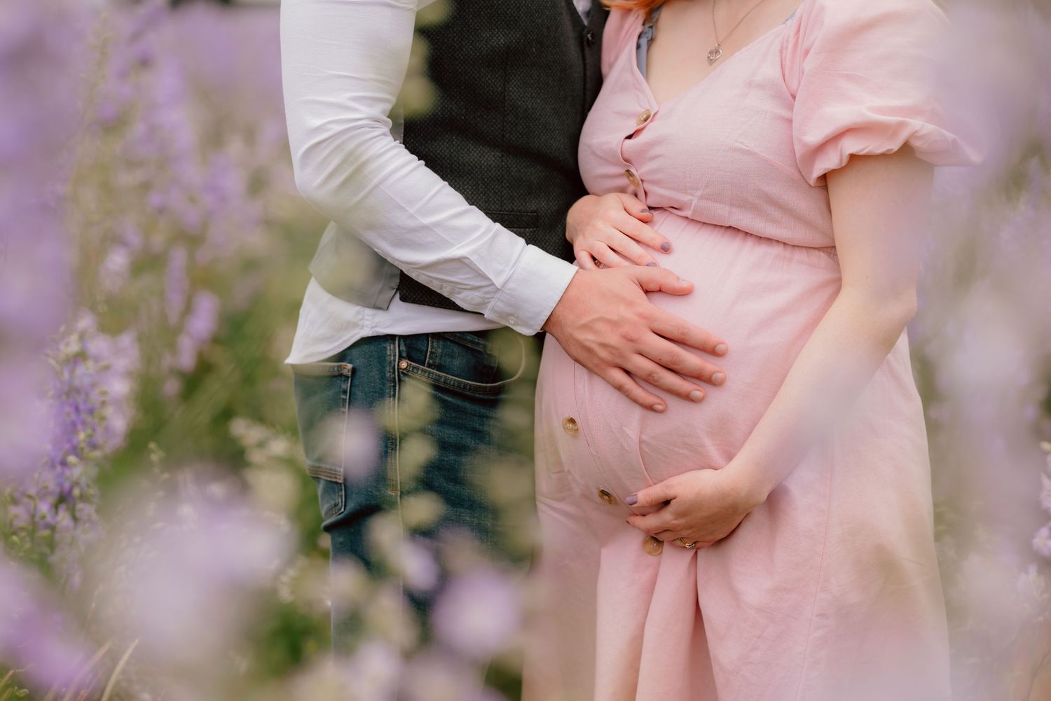 A tender pregnancy photo shows hands cradling a baby bump in a pink dress, surrounded by soft purple wildflowers and delphiniums.