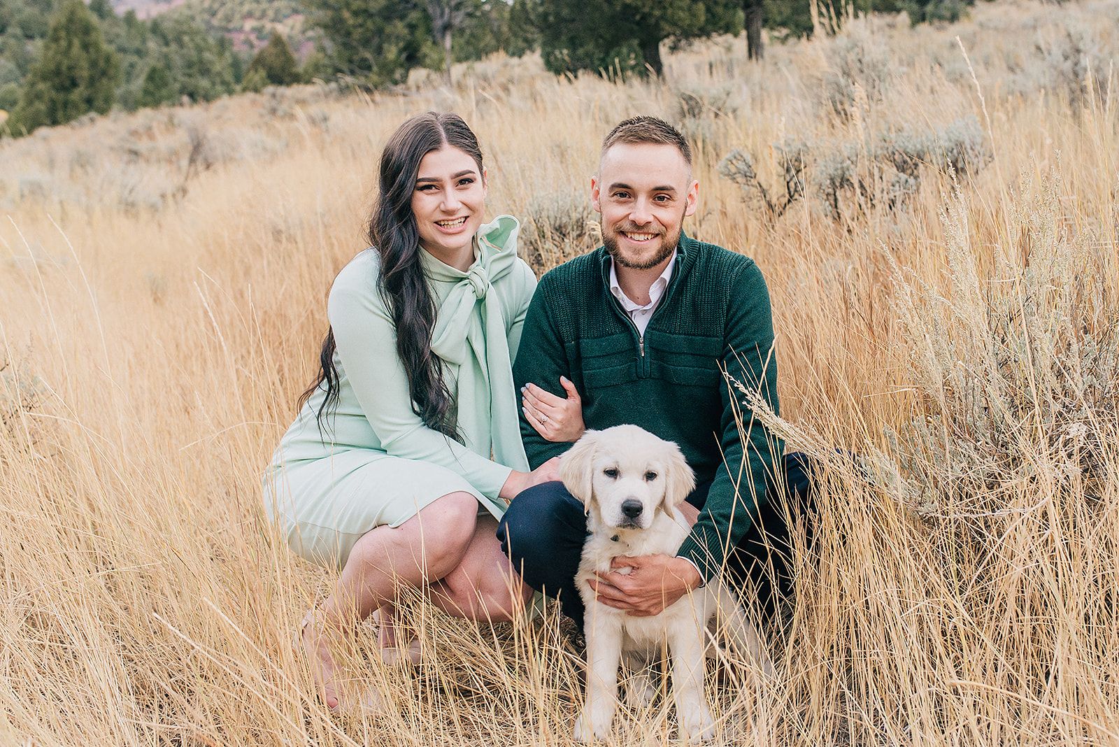 A couple sits in tall golden grass with their happy dog on a sunny day.