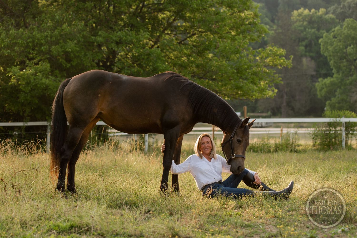 A peaceful scene of a dark horse grazing next to someone resting in a grassy field at sunset.