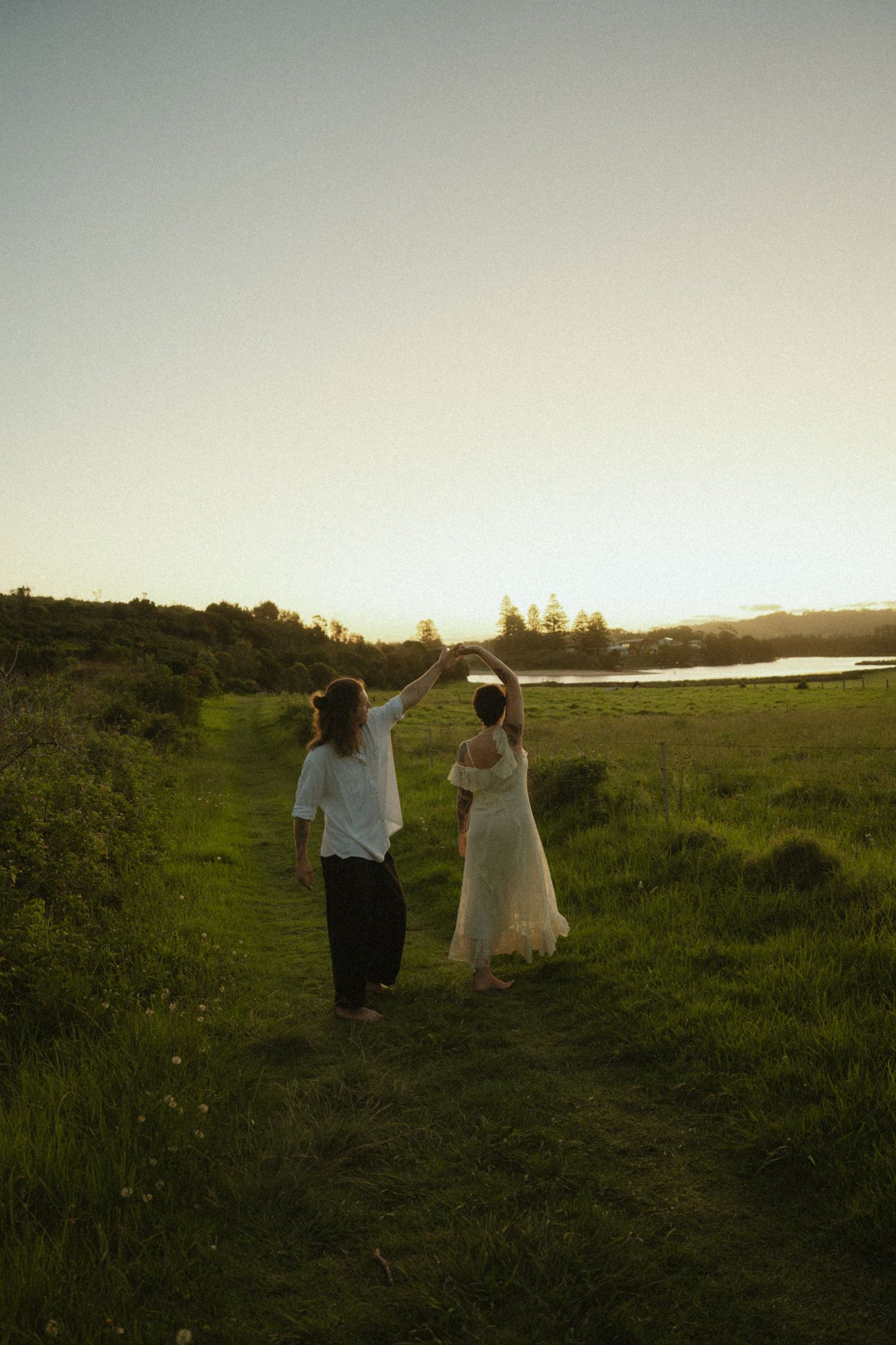 Two people walk hand in hand through a lush green meadow at sunset, their silhouettes outlined against the golden sky.