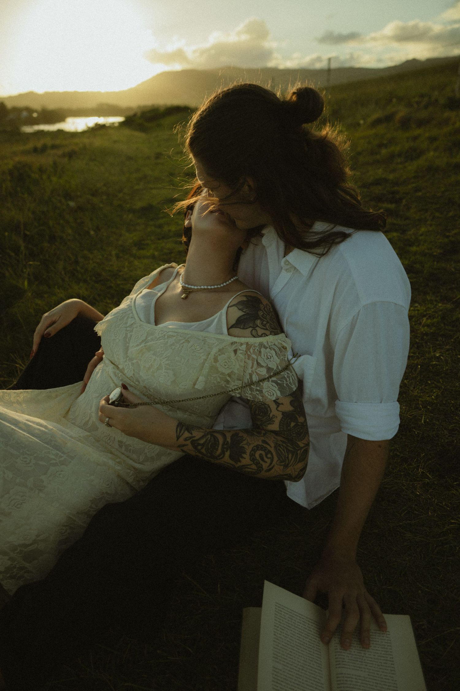 Romantic couple in vintage-style white outfits embrace on a grassy hillside at golden hour.