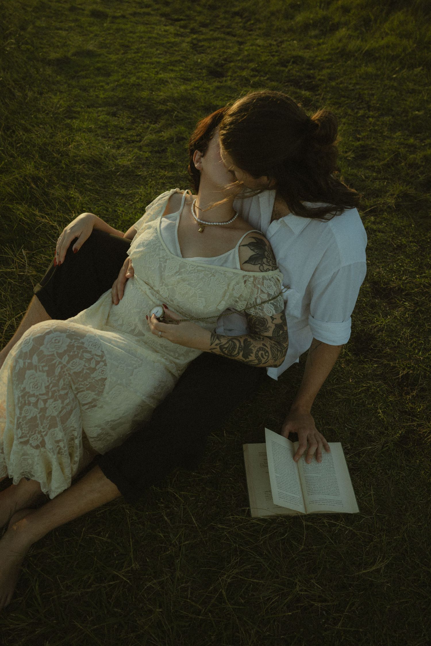 Two people in white outfits lie on grass reading books together in a romantic vintage-style scene.