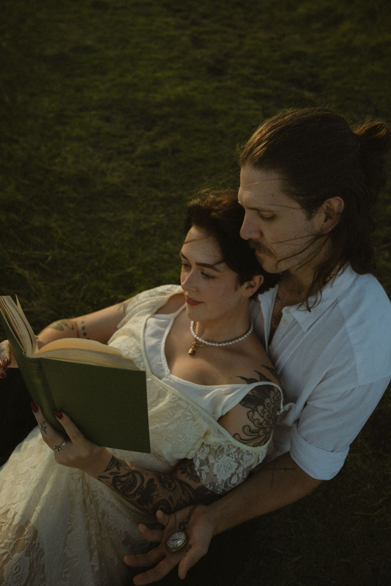 A couple in vintage-style clothing sits together on grass while reading a book in warm evening light.