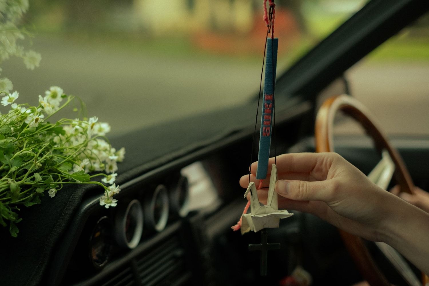 Close-up view of hands on steering wheel through car windshield with blooming flowers visible outside.