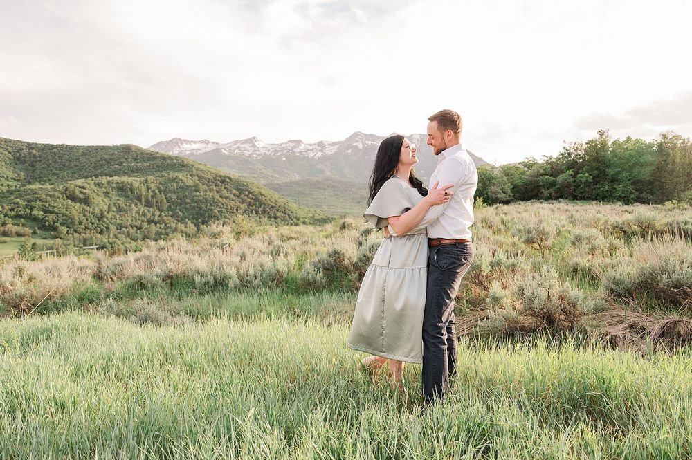 Couple embracing in evening light on grassy hillside with mountain vista behind them.