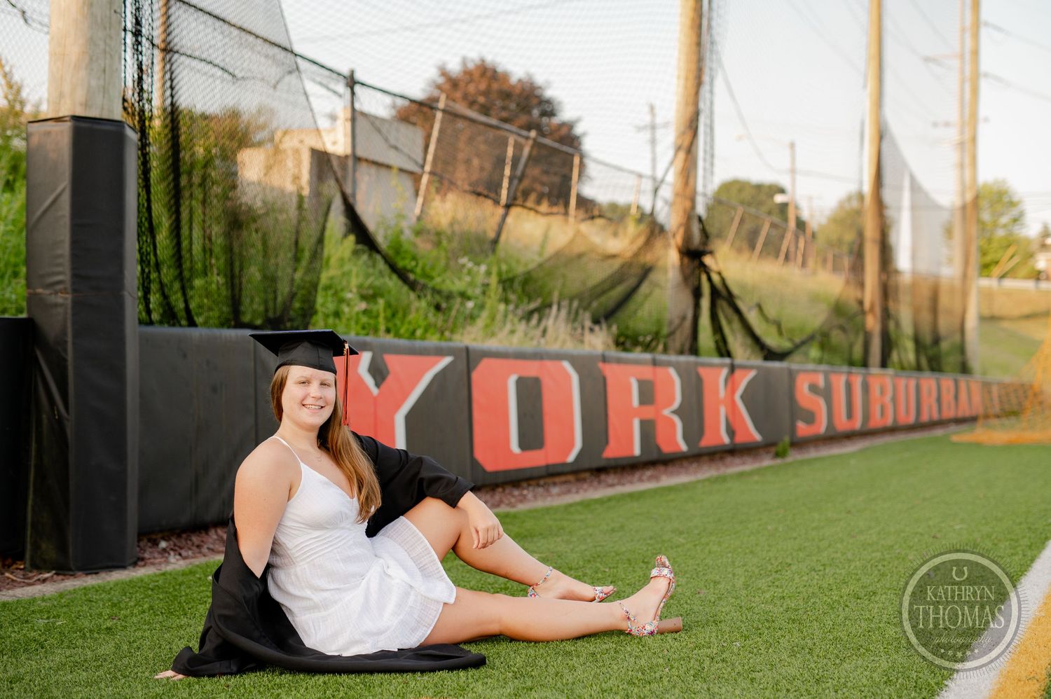 Graduate in white dress poses on sports field with York sign in background.