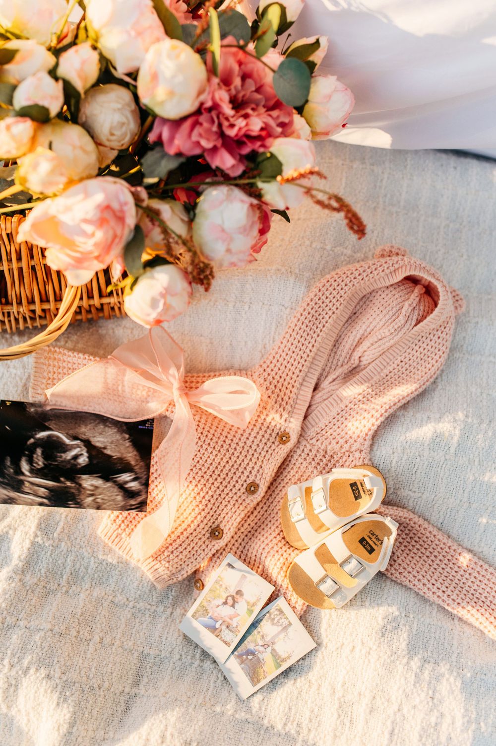 Pink knit baby clothes and tiny shoes arranged with delicate flowers for a maternity photoshoot.
