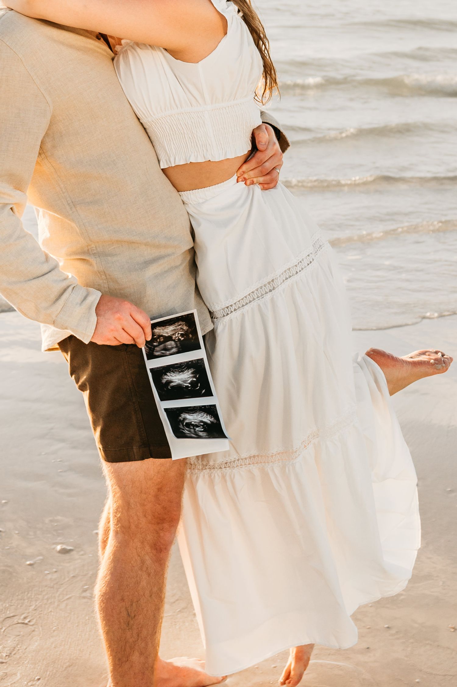Couple in white clothing embraces on beach while holding ultrasound photos during sunset pregnancy announcement photoshoot.