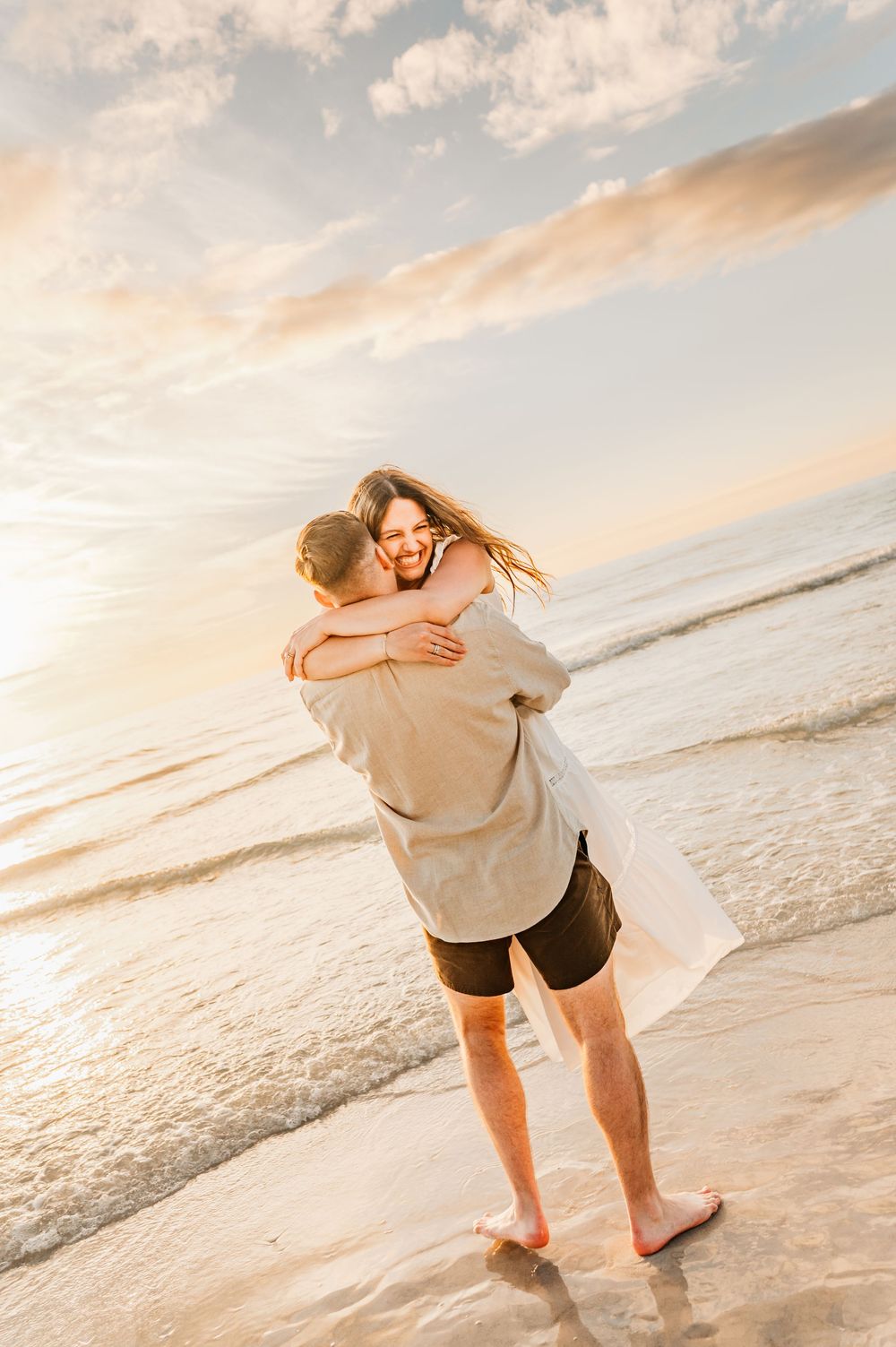 A couple embraces on a beach at sunset with soft golden light and ocean waves in the background.