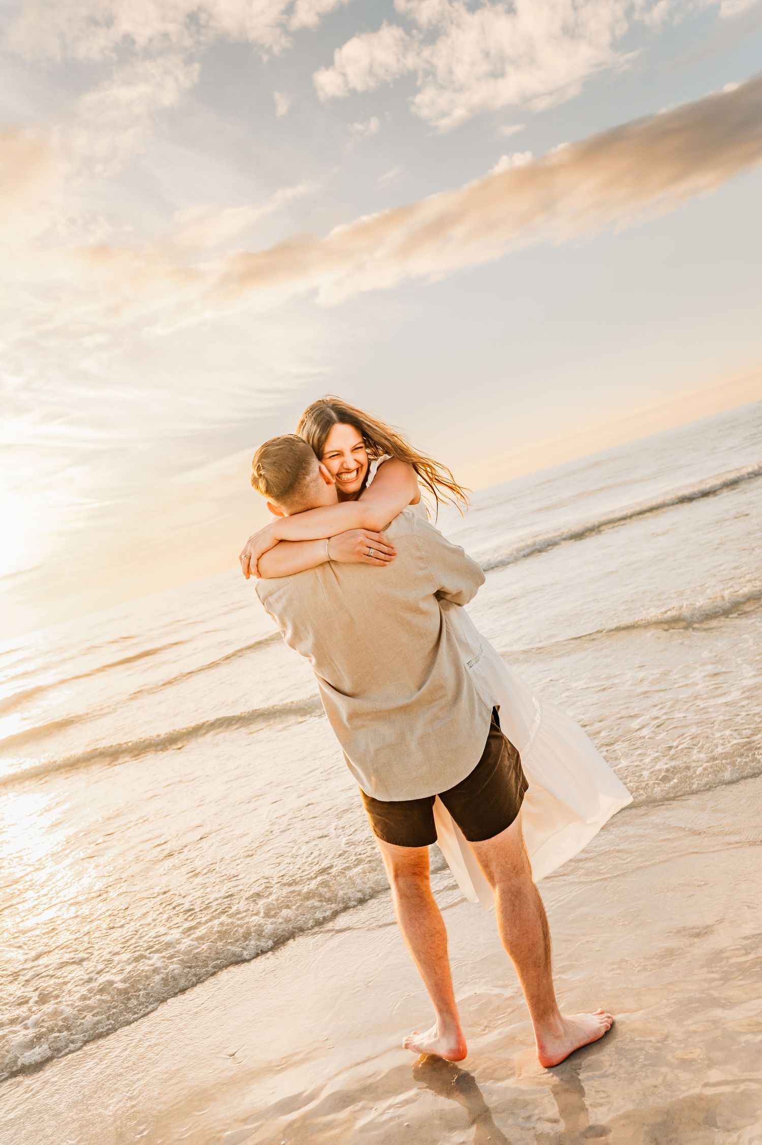 A couple embraces on a beach at sunset with soft golden light and ocean waves in the background.
