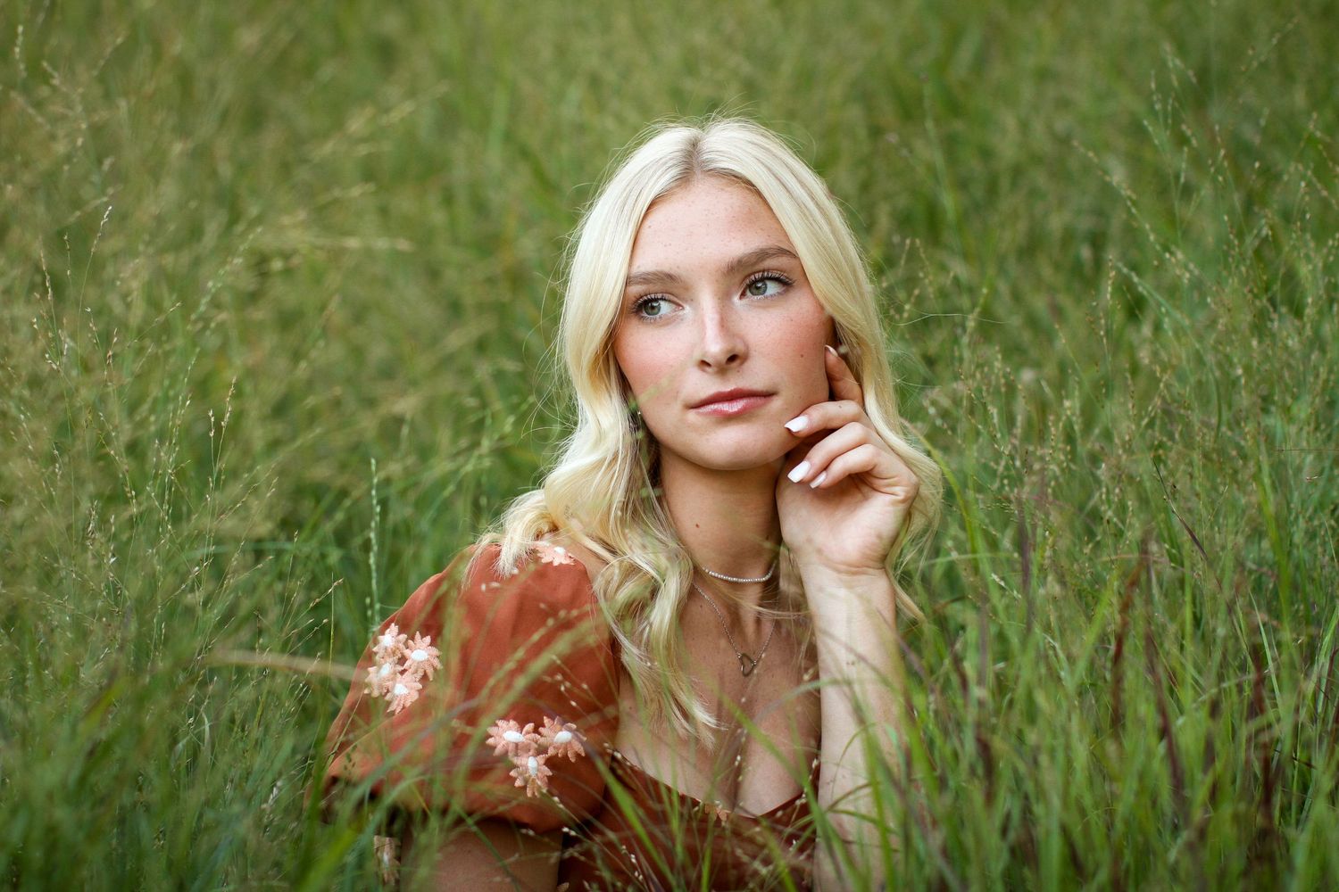 A blonde young woman surrounded by tall green grasses props her chin in her hand during her Kansas City senior photo session.