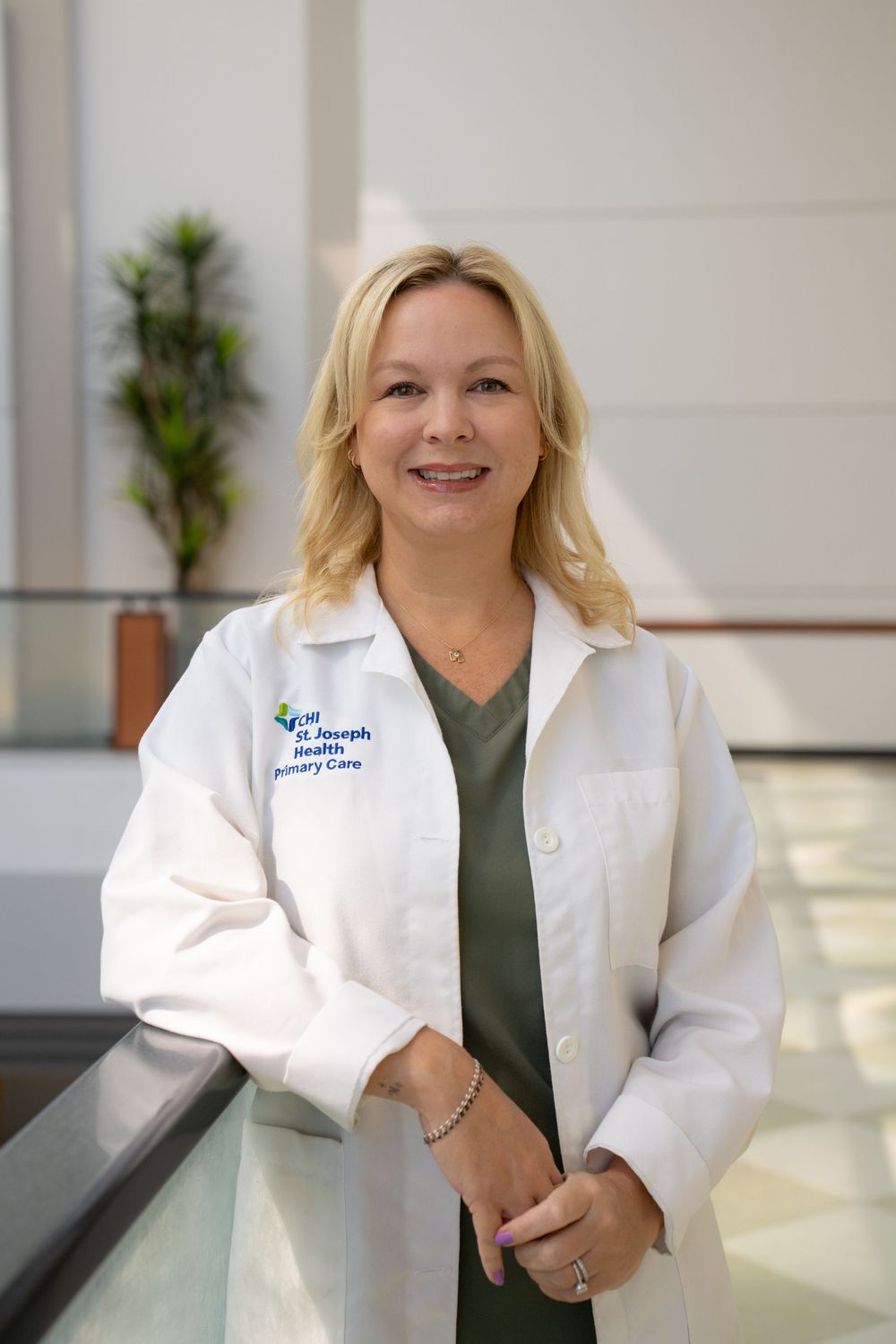 Healthcare professional in white lab coat and green scrubs smiling in a bright medical facility hallway.
