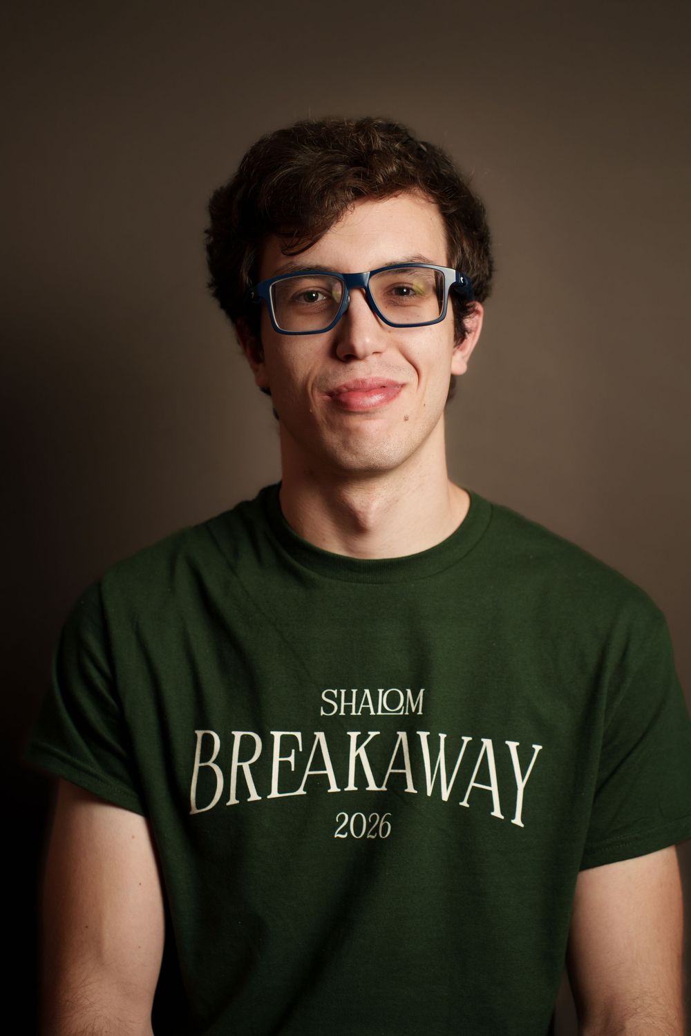 A young man with curly hair and glasses wearing a green t-shirt with the text 'Shalom Breakaway 2020'.