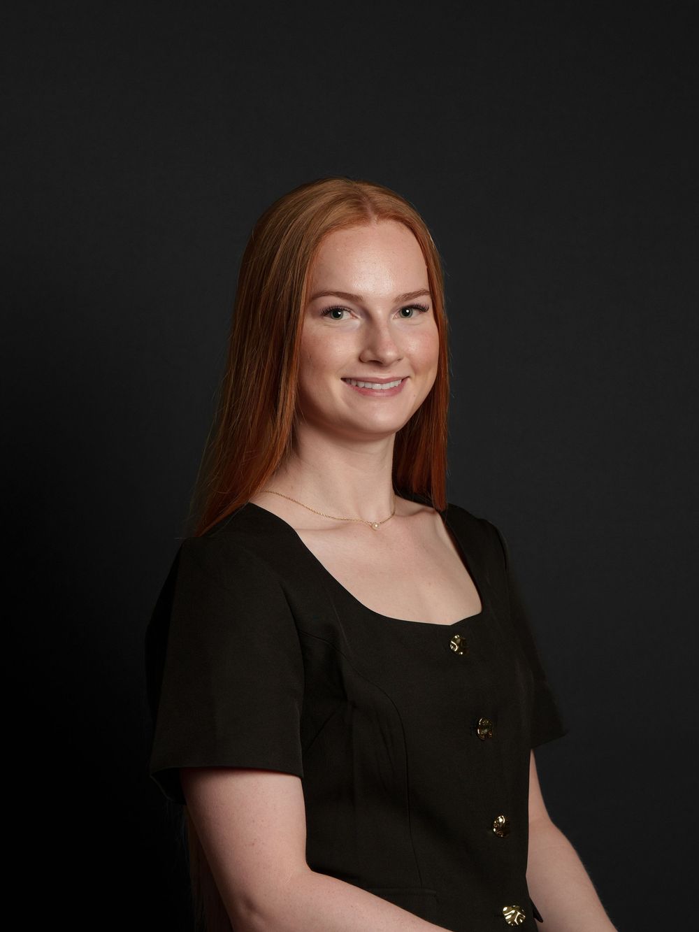 A smiling woman with long red hair wearing a black dress poses for a portrait against a dark background.