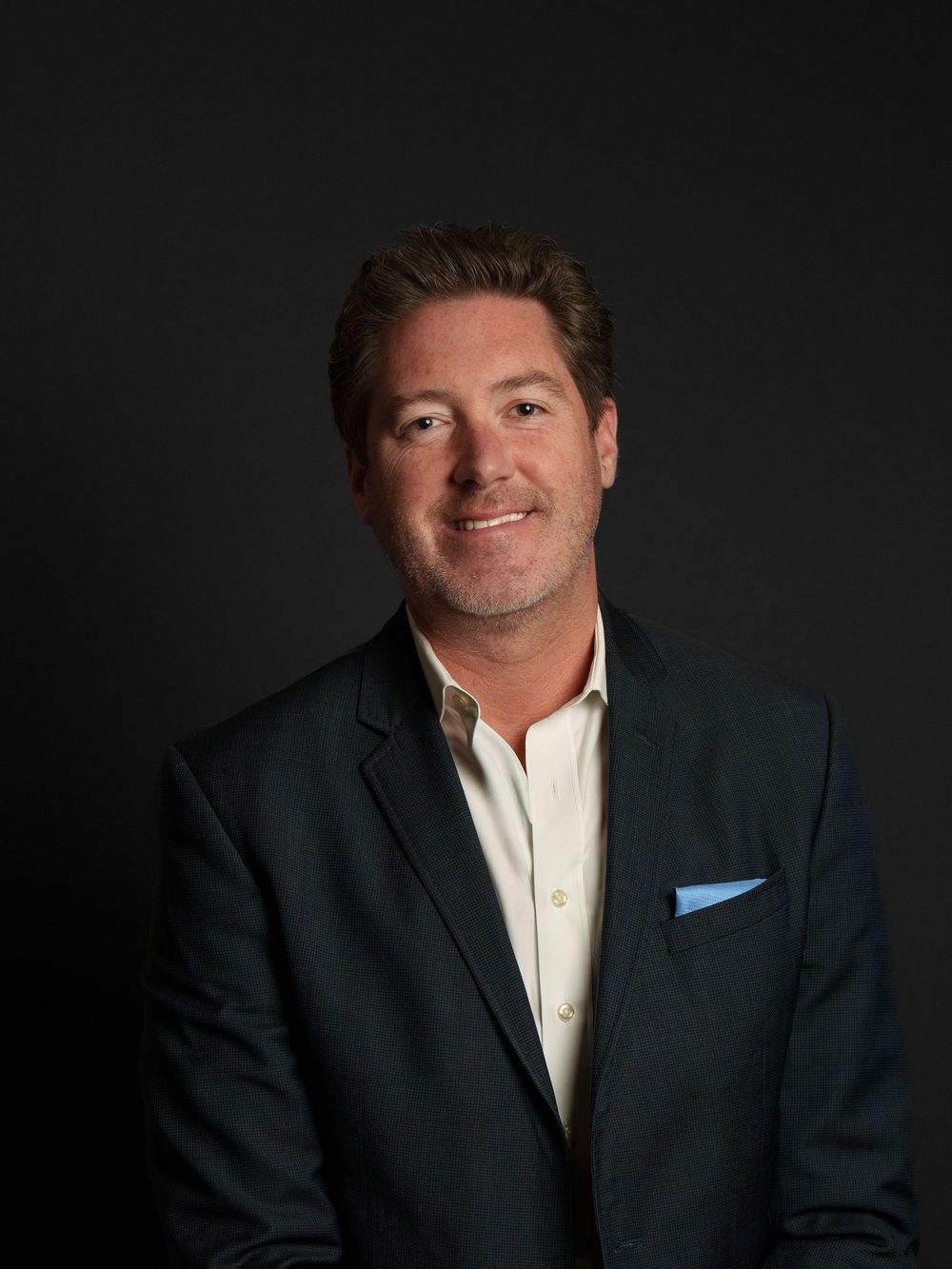 A middle-aged man with short brown hair wearing a black suit and blue pocket square smiles confidently in a studio portrait.