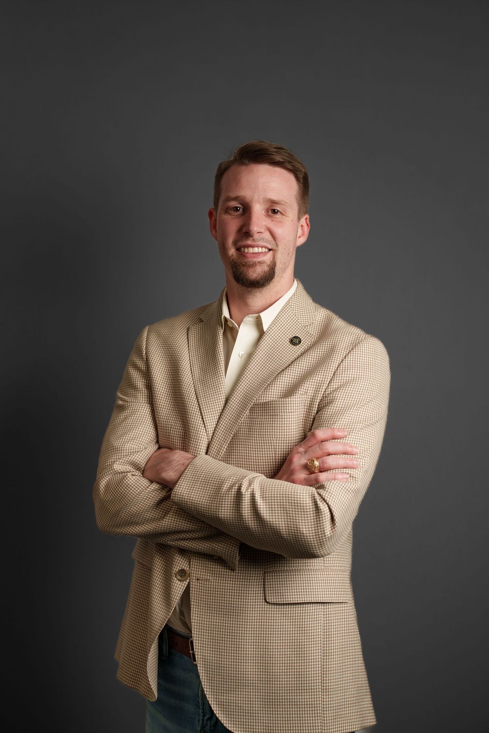 Smiling man in beige blazer with arms crossed poses confidently against gray background in professional headshot.