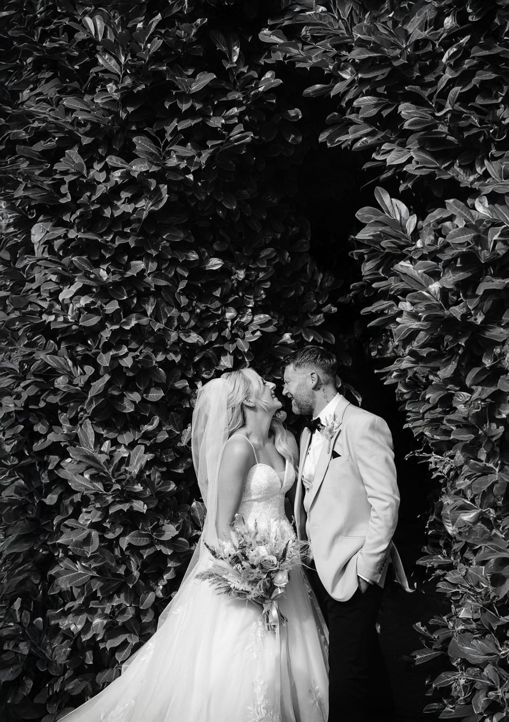 Bride and groom kissing under leafy green archway in black and white wedding portrait.
